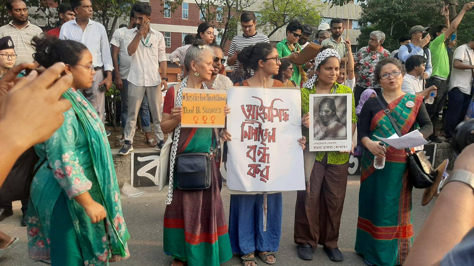 Women and activists gather with signs at a public demonstration, demanding justice and freedom from silence.